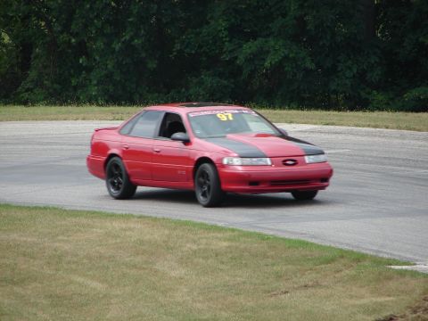 1993 Ford Taurus - A red sedan with black hood stripes is driving on a race track, displaying the number 97.