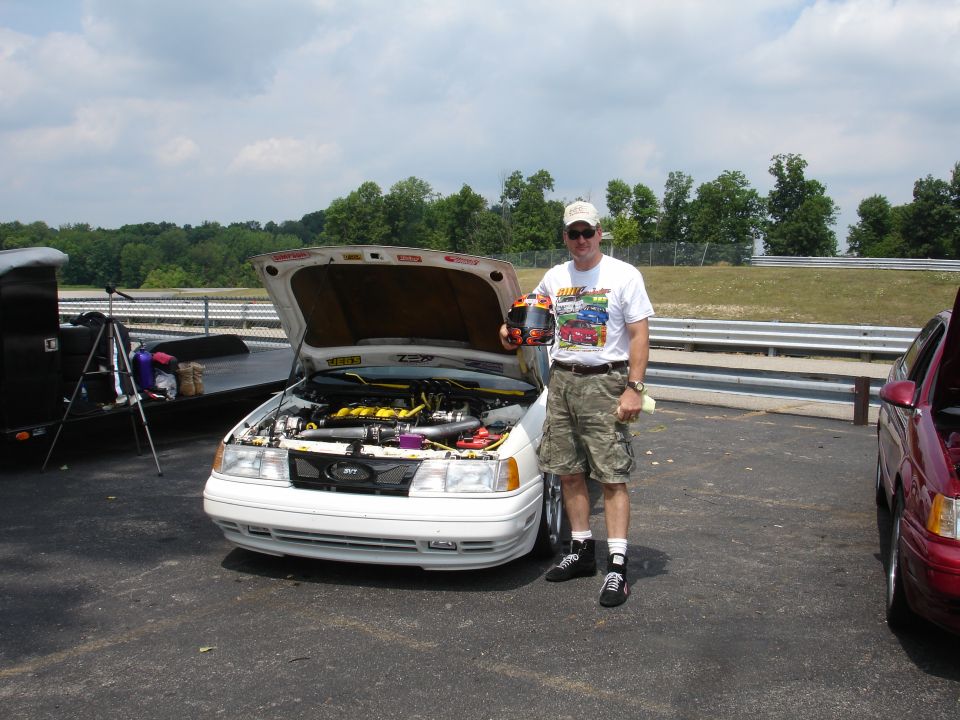 Man holding helmet stands next to white car with hood open. Another red car is parked to the right