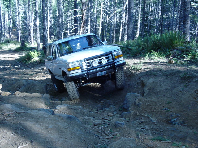 White Ford Bronco with auxiliary lights crawling a rocky rutted trail in dense forest