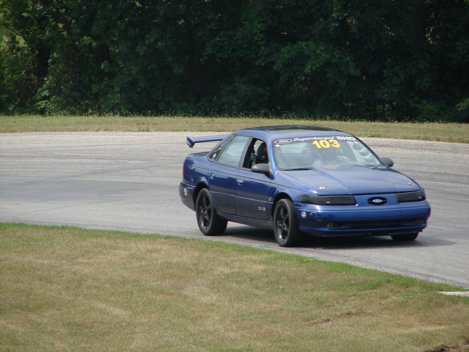 Blue 1993 Ford Taurus with a spoiler and number 103 on the windshield driving on a race track