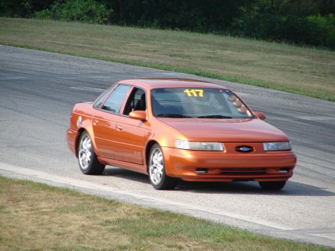 1993 Ford Taurus - A bright orange sedan navigates a turn on a paved race track with grassy shoulders.