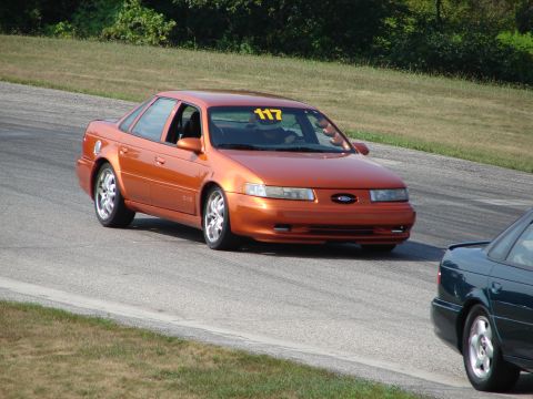 1993 Ford Taurus - An orange 1993 Ford Taurus, identified by its body style and race number 117, navigates a turn on a road course.