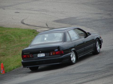 1993 Ford Taurus - A black 1993 Ford Taurus sedan navigates a turn on a race track, with tire marks visible on the asphalt.