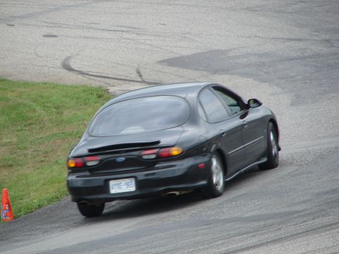 1993 Ford Taurus - A black sedan with a distinctive oval rear window navigates a road course, leaving tire marks on the asphalt.