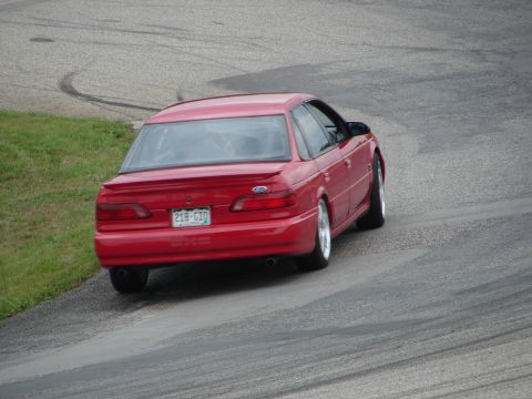 1993 Ford Taurus - A red 1993 Ford Taurus sedan is seen from the rear on a road course, with tire marks visible on the asphalt.