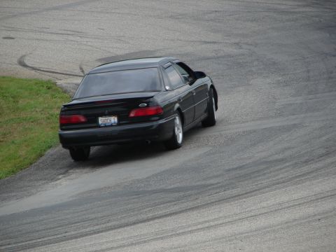 1993 Ford Taurus - A black 1993 Ford Taurus is captured on a race track, with tire marks visible on the asphalt.