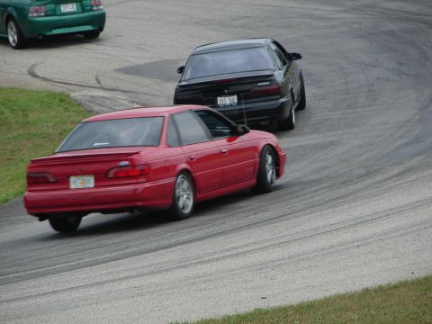 1993 Ford Taurus - A red 1993 Ford Taurus sedan navigates a turn on a road course, with other vehicles visible in the background.