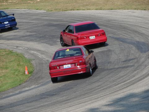 1993 Ford Taurus - Two red sedans navigate a turn on a race track, with one displaying a racing number.