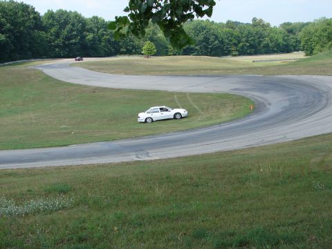 1993 Ford Taurus - A white sedan is parked on the grass beside a winding race track, with another vehicle visible in the distance.