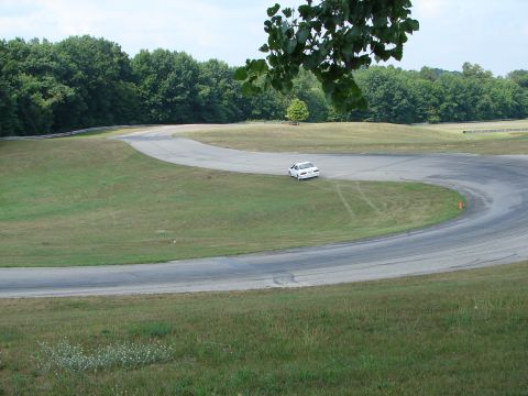 1993 Ford Taurus - A white sedan navigates a turn on a paved race track surrounded by grassy areas and trees.