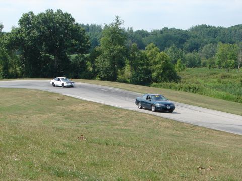 1993 Ford Taurus - A green sedan with racing number 64 on its windshield navigates a turn on a road course, with another white car visible in the distance.