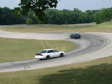 1993 Ford Taurus - Two cars drift around a turn on a paved road course with grassy verges and trees.