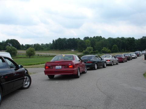 1993 Ford Taurus - A line of cars, including a red 1993 Ford Taurus, waits on a paved surface with trees in the background.