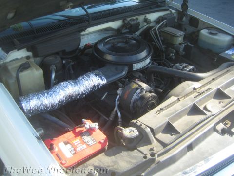 1990 Ford Bronco - Open engine bay of an older vehicle showing a round air cleaner, a foil-wrapped intake hose, and a red battery box.