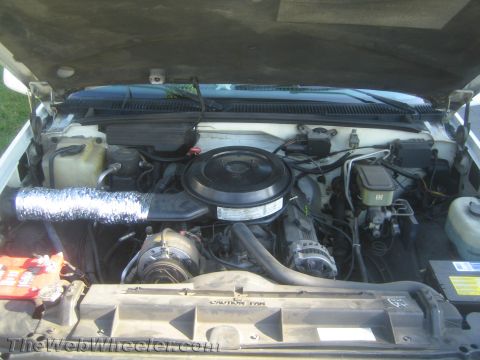 1990 Ford Bronco - Open engine compartment of an older vehicle with a round air cleaner, foil-wrapped intake hose, and a fan shroud marked Caution Fan.
