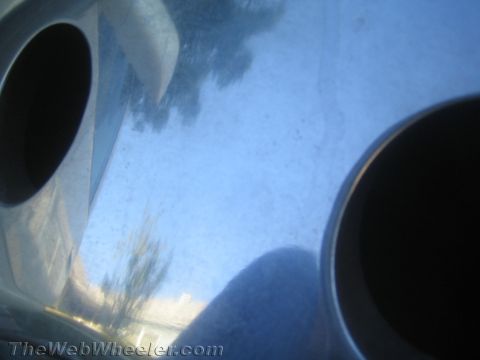 1990 Ford Bronco - Blurry overexposed close-up of a polished metal wheel face showing dark cutout holes and faint tree and sky reflections.