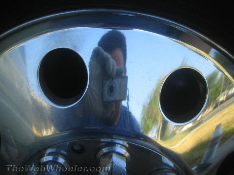 1990 Ford Bronco - Polished chrome wheel face with two dark holes reflecting the photographer holding a camera and wearing a glove, with grass behind.