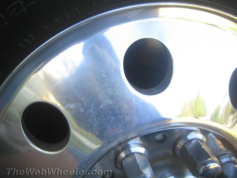 1990 Ford Bronco - Tight close-up of a polished aluminum wheel showing oval cutout holes, the tire shoulder, and chrome lug nuts at the hub.