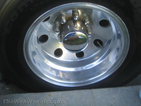 1990 Ford Bronco - Front view of a polished aluminum wheel with oval cutouts, a chrome center cap and lug nuts, mounted on a tire on the ground.