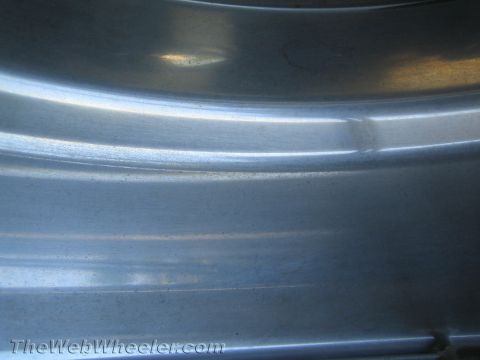 1990 Ford Bronco - Extreme close-up of a curved brushed metal surface, likely a wheel rim, showing ridges and soft light reflections.