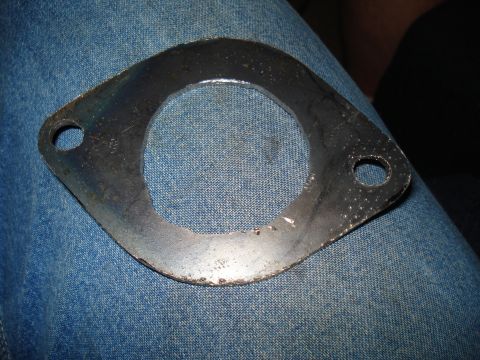 1967 Ford F-250 - A metal exhaust flange gasket with two bolt holes is shown resting on blue denim fabric.