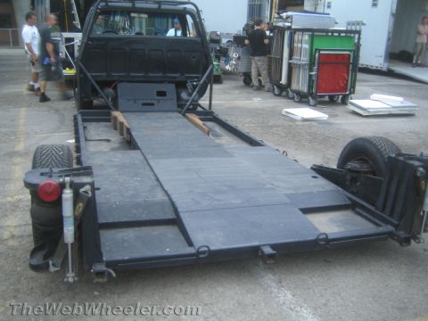 1990 Ford Bronco - black flatbed ramp truck with its bed tilted to the ground, parked in a busy equipment lot with people and carts.