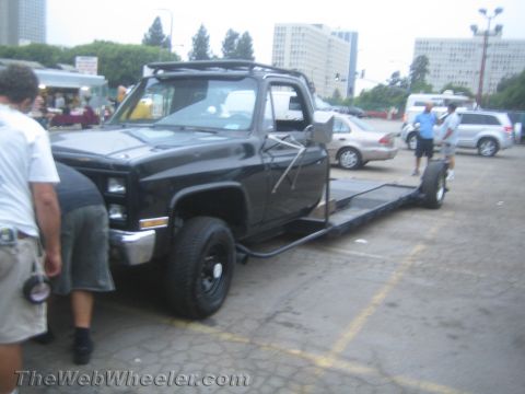 1990 Ford Bronco - black squarebody Chevrolet pickup fitted with a long flatbed ramp, people standing beside it in a city parking lot.