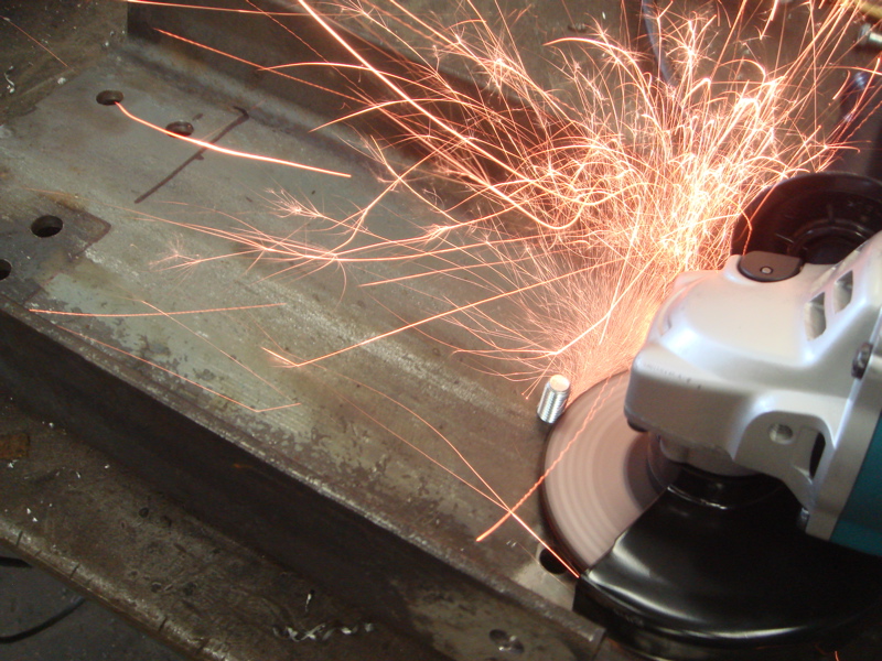 Angle grinder sparks fly as it cuts a metal beam with holes drilled in it