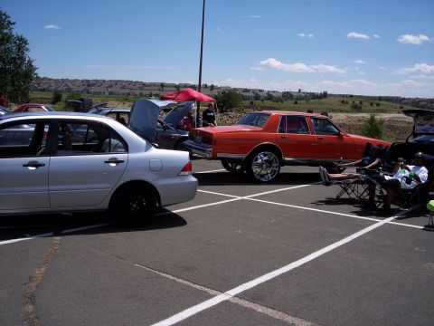 1972 Ford Ranchero - An orange sedan with large chrome rims is parked at an outdoor event, with people relaxing in chairs nearby.
