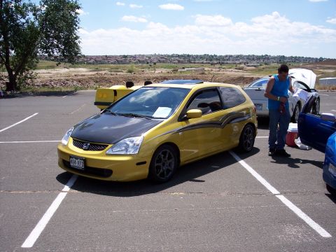 1972 Ford Ranchero - A yellow hatchback with a carbon fiber hood and racing stripes is parked in a lot.