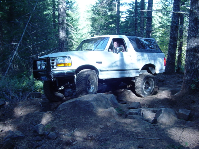 White lifted Ford Bronco with brush guard flexing over a large rock on a forest trail