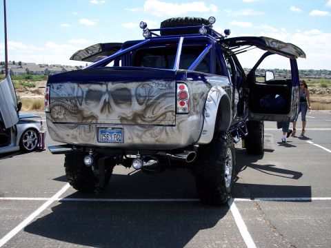 1972 Ford Ranchero - A heavily customized pickup truck with a skull airbrushed on the tailgate and a roll cage is parked next to a car with unique doors.