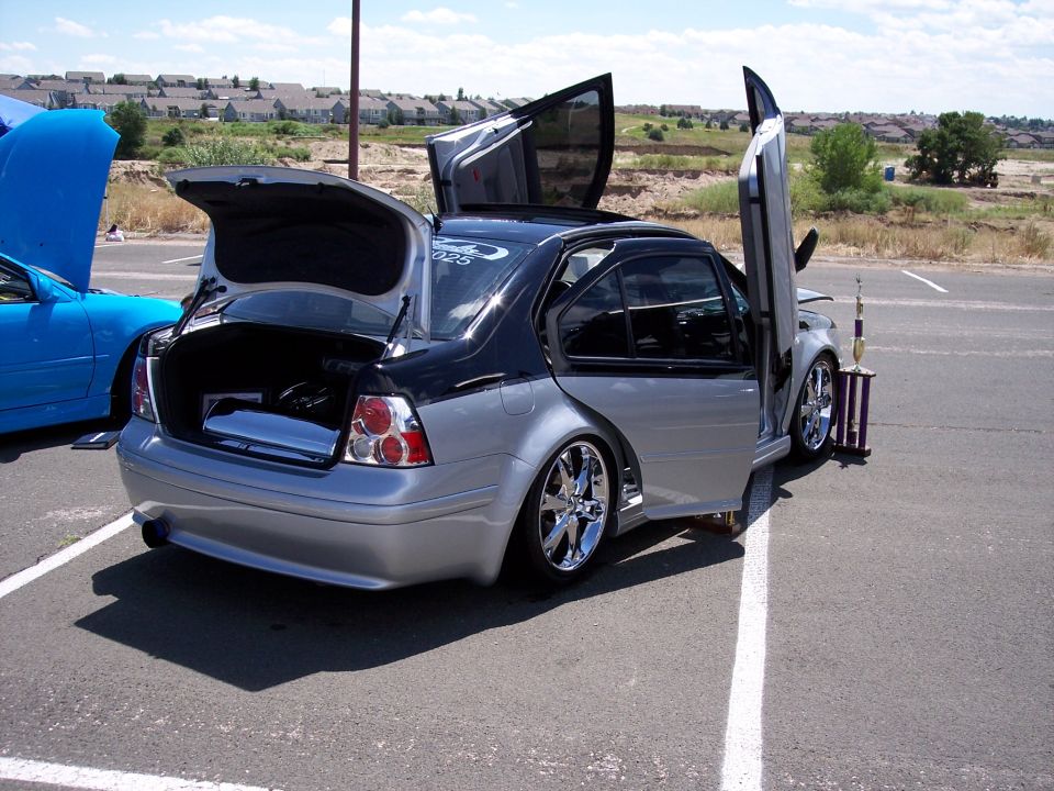 Silver and black sedan with gullwing doors open, trunk open, and a trophy beside it. Blue car with hood open in background