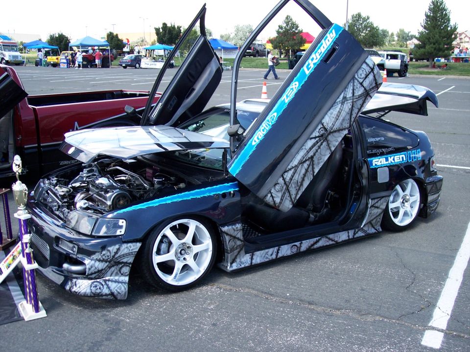 Customized black Honda CRX with lambo doors open, engine exposed, and custom paint job. Trophy in foreground
