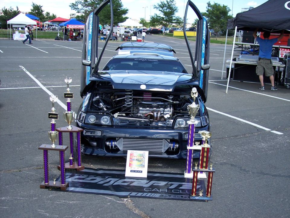Dark blue coupe with lambo doors open, hood up, and trophies in front