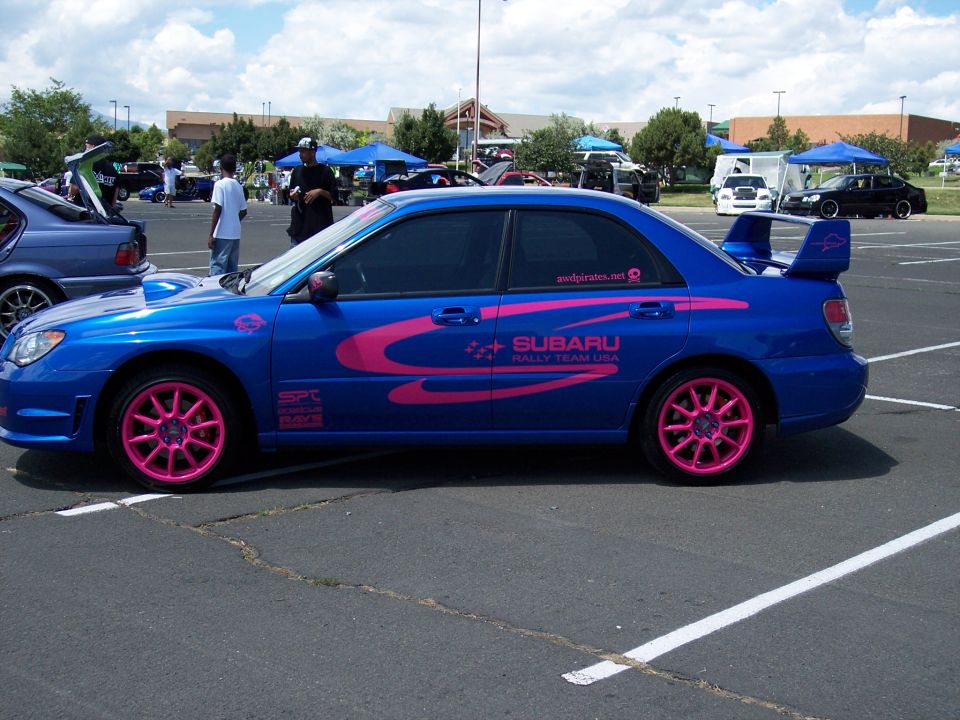 Blue Subaru WRX STI with pink racing stripes and pink wheels, parked in a lot. "SUBARU RALLY TEAM USA" text visible