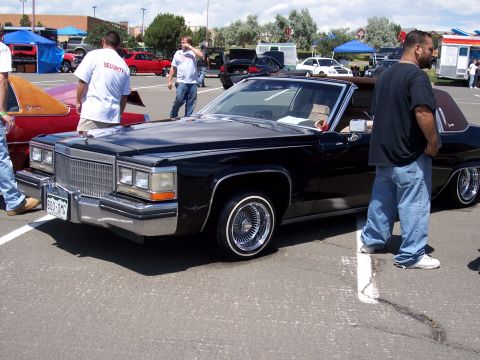 1972 Ford Ranchero - A black Cadillac convertible is parked in a lot, with several people milling about. The license plate reads 600-OMG.