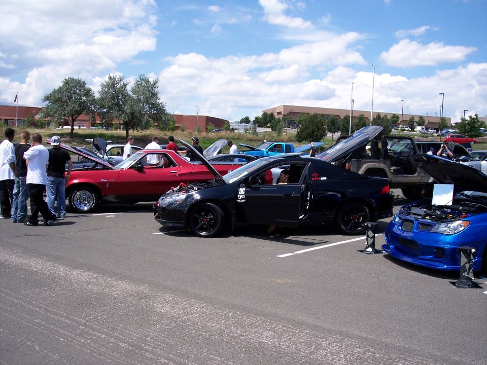 Several cars are parked in a lot with hoods open, people are milling around
