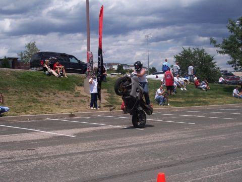1972 Ford Ranchero - A motorcycle rider performs a wheelie in a parking lot while onlookers watch from a nearby hill.