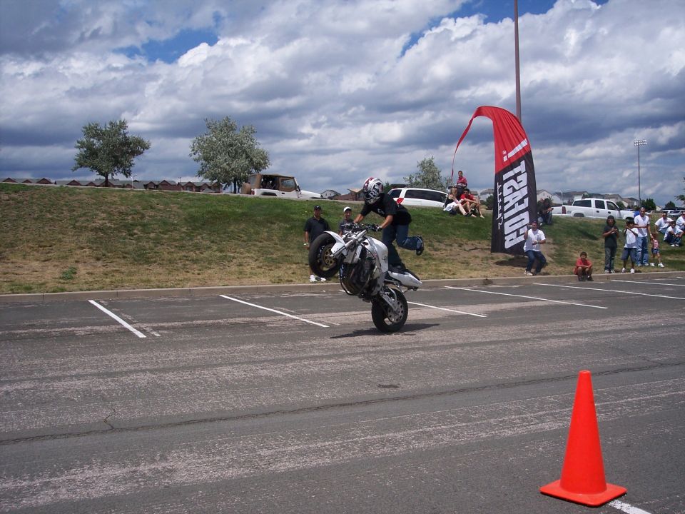White motorcycle performing a wheelie in a parking lot, with spectators and a large red banner in the background