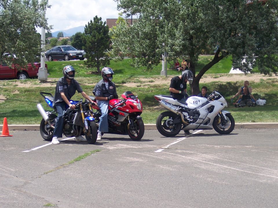 Three sport motorcycles parked in a lot, two with riders. A white motorcycle has 'BI!' on its side