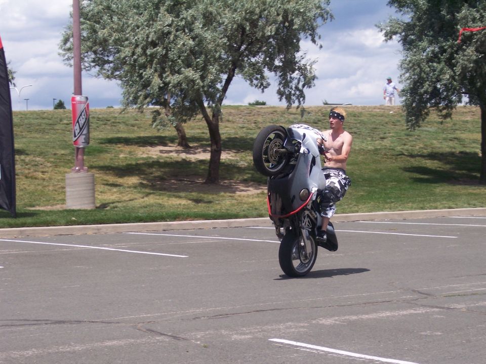 Guy on a motorcycle doing a wheelie in a parking lot. He's wearing a bandana and camo pants