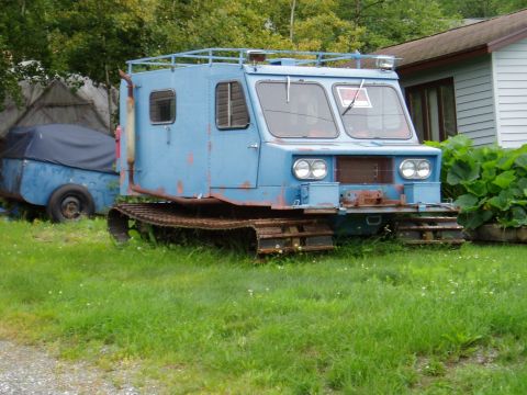 2000 Ford E350 - A blue tracked vehicle sits in a grassy area next to a covered trailer and a house.