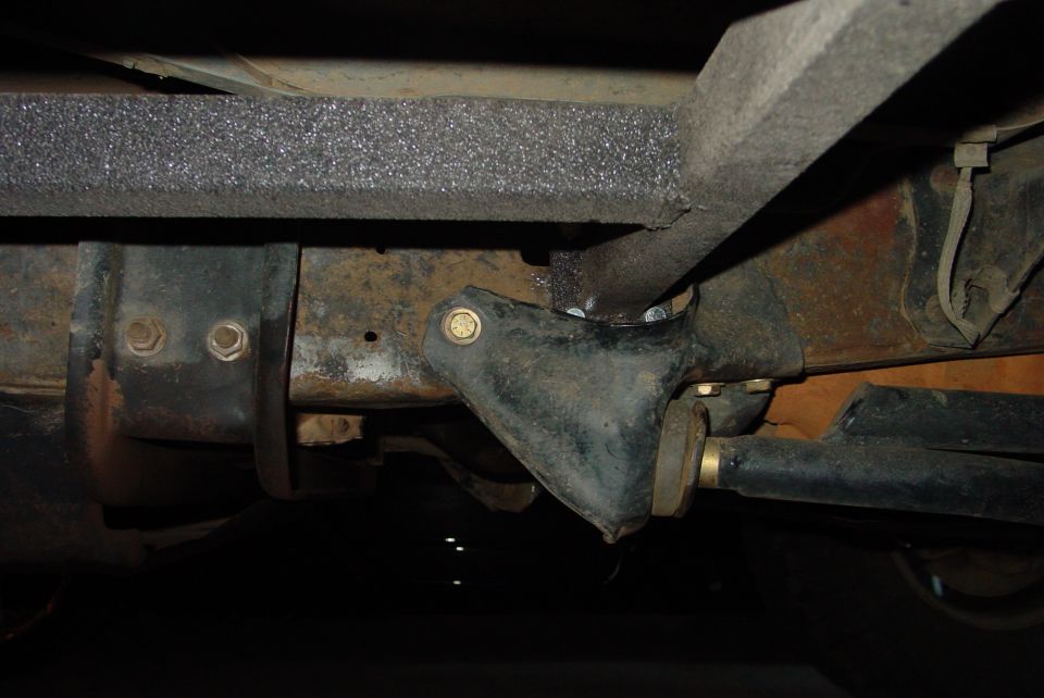Underside view of vehicle frame with rust and aftermarket sliders attached. Visible bolts and suspension parts