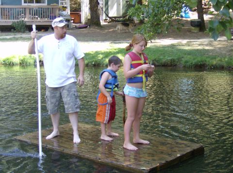 1986 Ford Bronco - A man and two children stand on a raft in the water, with a house and trees visible behind them.