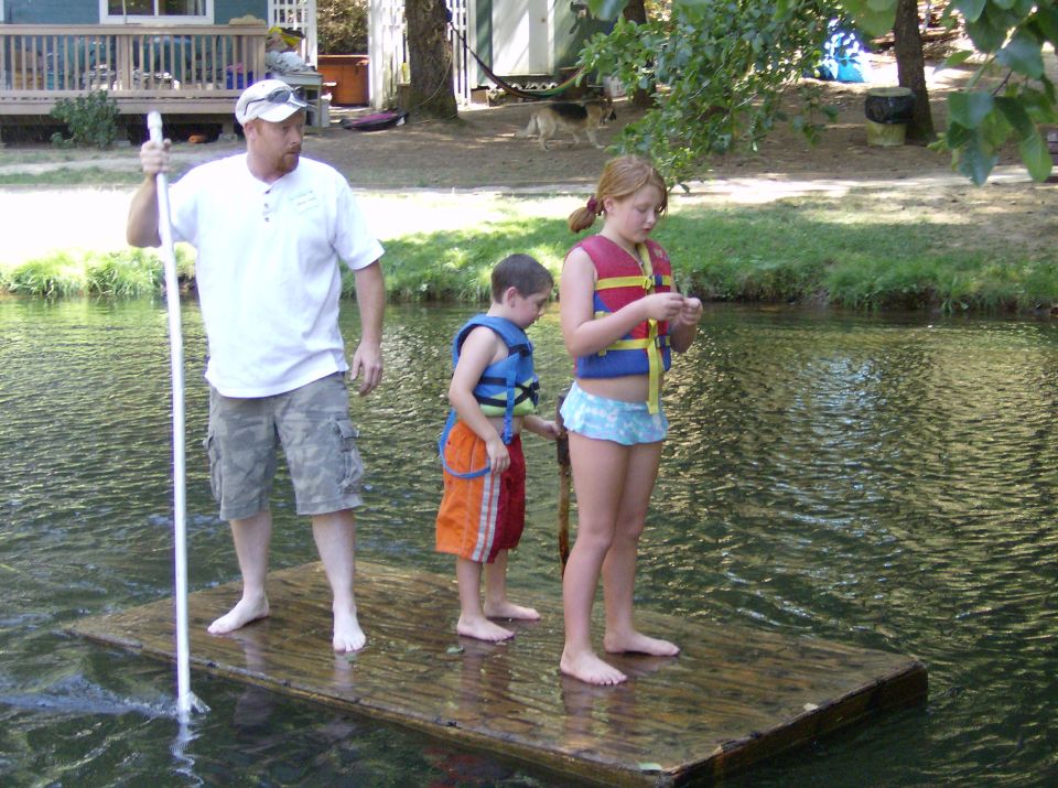 Man, boy, and girl on a wooden raft in a body of water, with a house and trees in the background