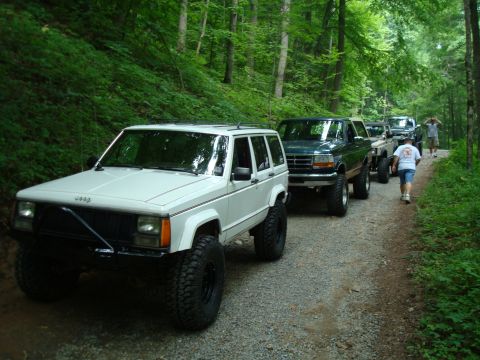 1995 Ford Bronco - A convoy of off-road vehicles, including a white Jeep Cherokee, travels down a wooded gravel path.