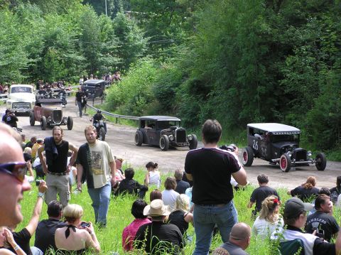 1978 Ford Bronco - A procession of vintage hot rod cars drives down a dirt road, observed by a crowd of onlookers.