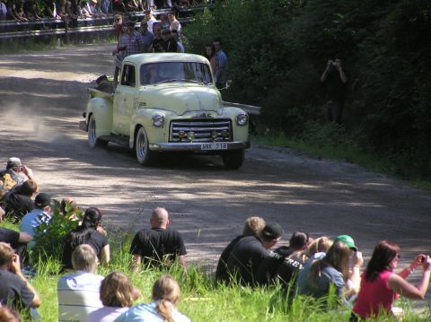 1978 Ford Bronco - A classic yellow GMC pickup truck drives on a dirt track as spectators watch from the roadside.
