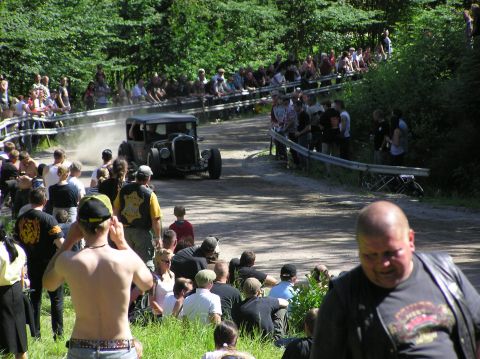 1978 Ford Bronco - A crowd watches a vintage hot rod race down a dusty track lined with trees and barriers.